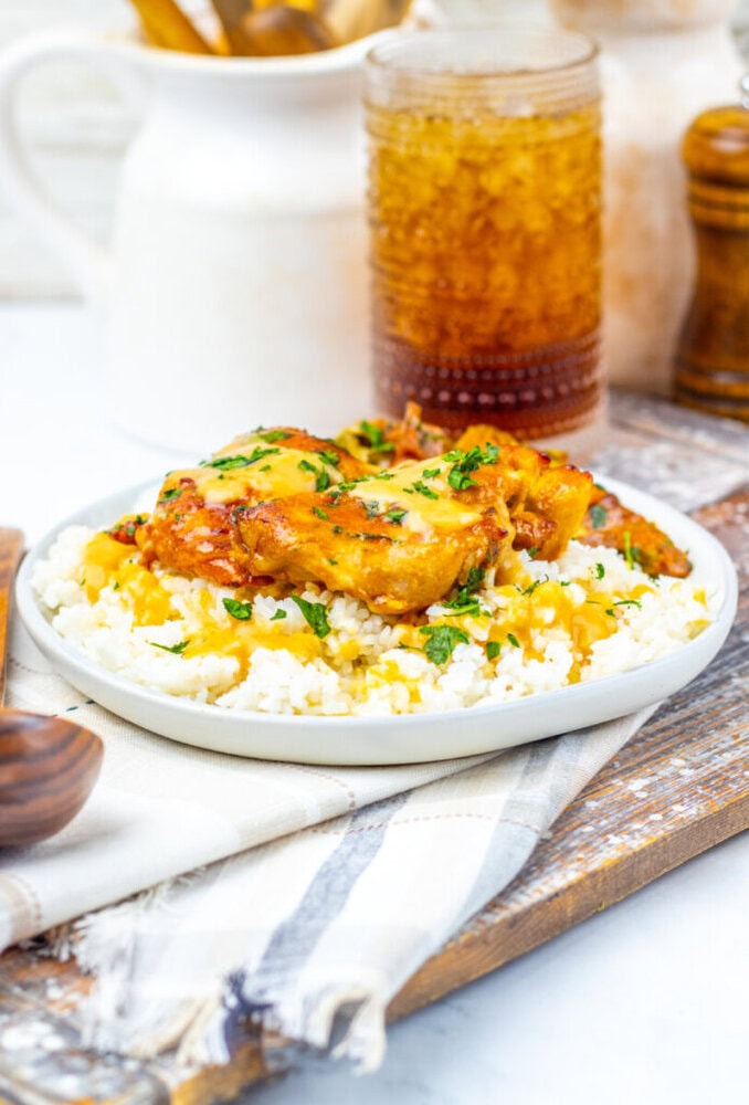 A plate of rice topped with saucy chicken and herbs sits on a cloth napkin, with a glass of iced drink and condiments in the background.