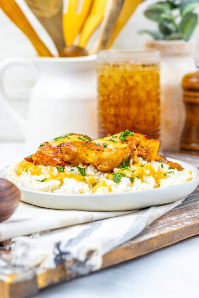 A plate of cooked chicken in sauce served over rice, garnished with herbs, sits on a wooden board with a glass of iced drink and kitchen utensils in the background.