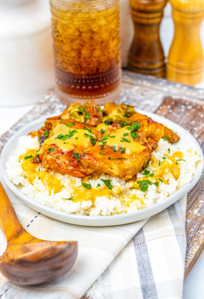 A plate of white rice topped with cheesy baked chicken and herbs, served with a wooden spoon; a drink and condiments are in the background.