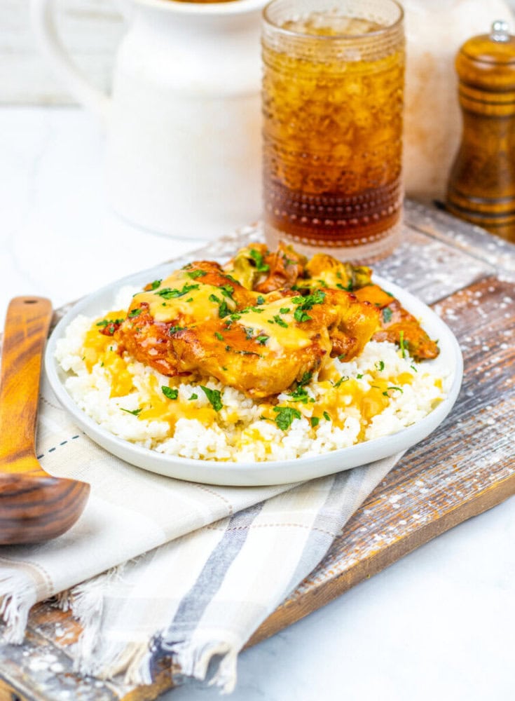 A plate of white rice topped with saucy chicken pieces and chopped herbs, placed on a napkin with a wooden spoon beside it and a glass of iced drink in the background.