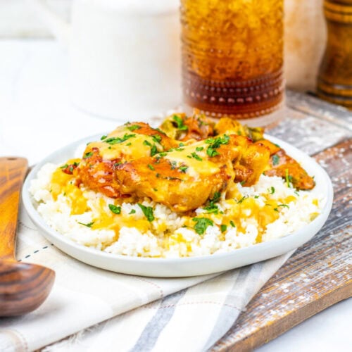 A plate of cooked chicken with sauce and herbs served over white rice, next to a wooden spoon and beverage, on a wooden board with salt and pepper shakers in the background.
