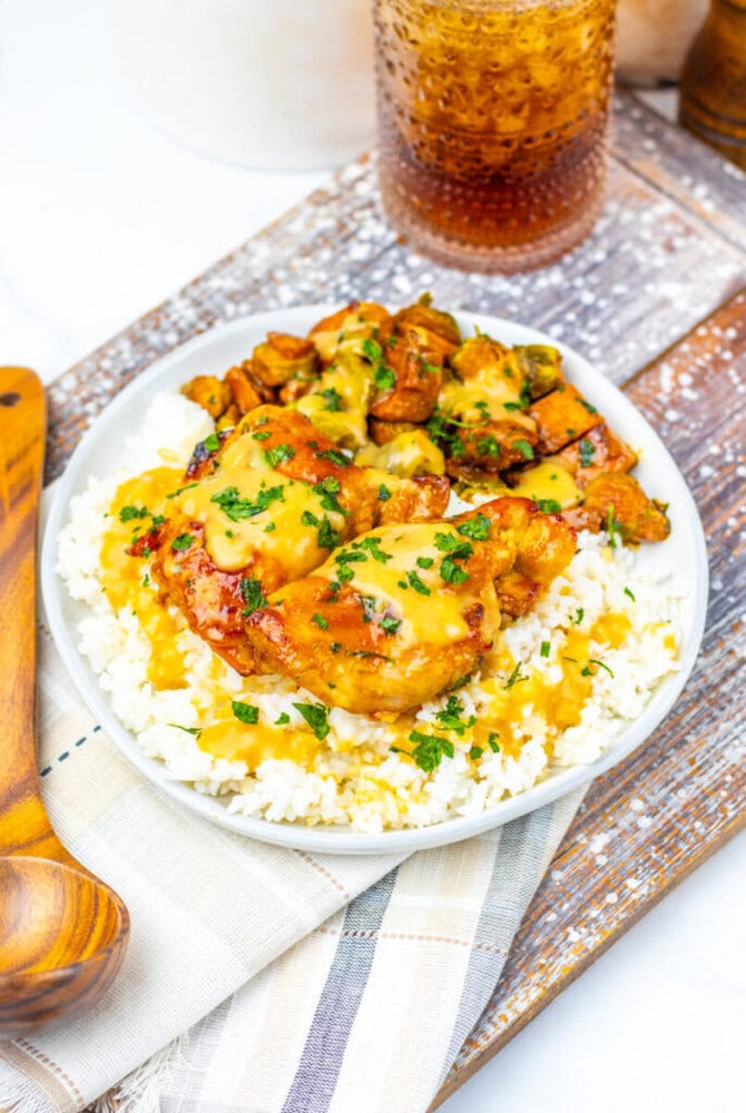 A plate of white rice topped with pieces of saucy grilled chicken, garnished with chopped herbs, next to a glass of iced drink on a wooden tray.