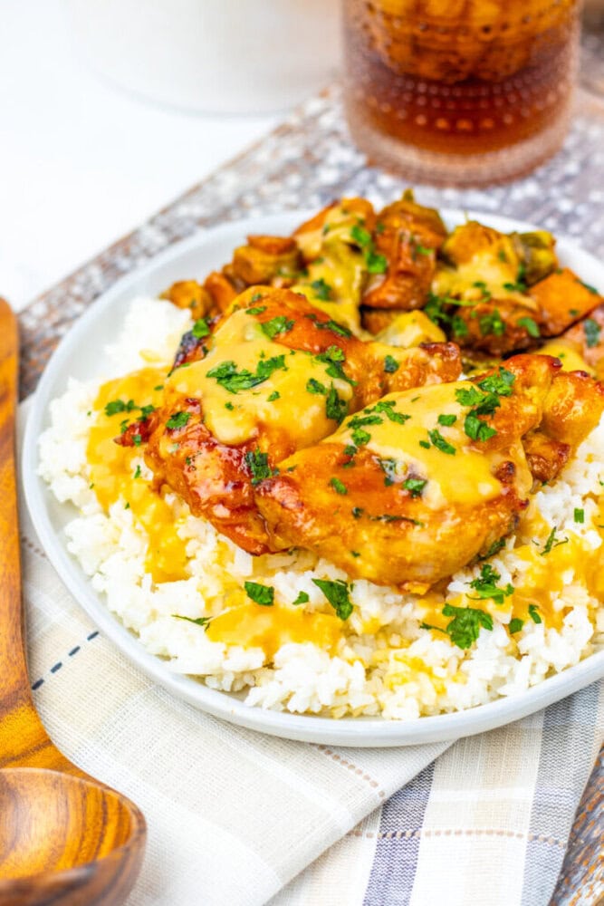A plate of white rice topped with pieces of chicken in a yellow sauce, garnished with chopped herbs, sits on a cloth napkin beside a wooden spoon and a glass of soda.