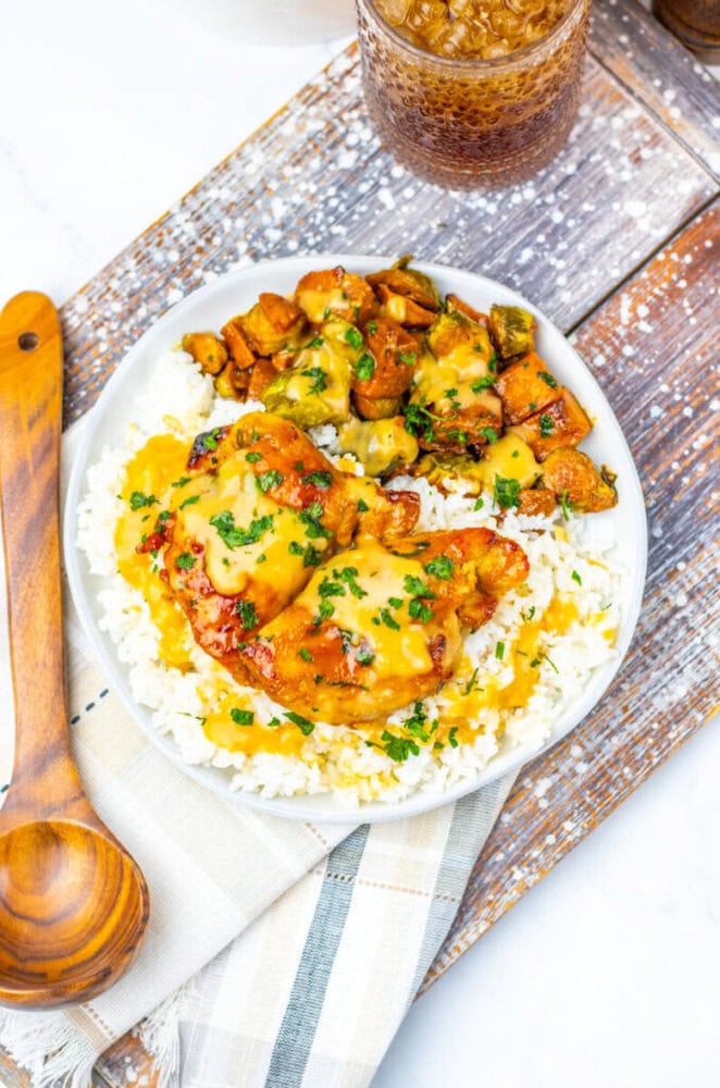 A plate of white rice topped with saucy chicken thighs and diced vegetables, garnished with chopped herbs, next to a wooden spoon and a glass of iced drink.