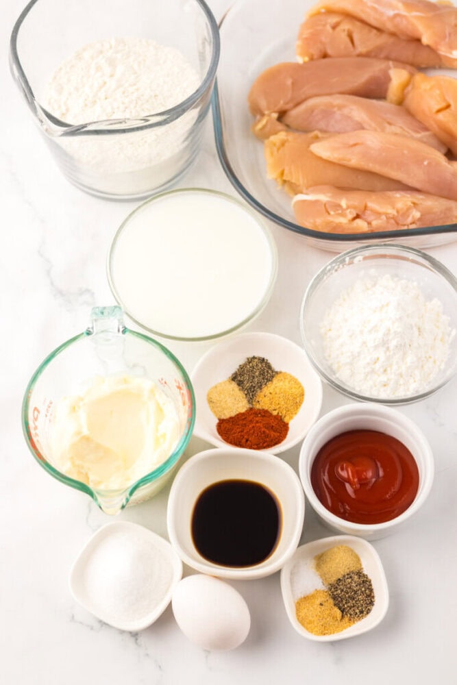 Various ingredients for making chicken strips are arranged on a white surface, including raw chicken, flour, milk, mayonnaise, spices, egg, ketchup, soy sauce, and cornstarch.