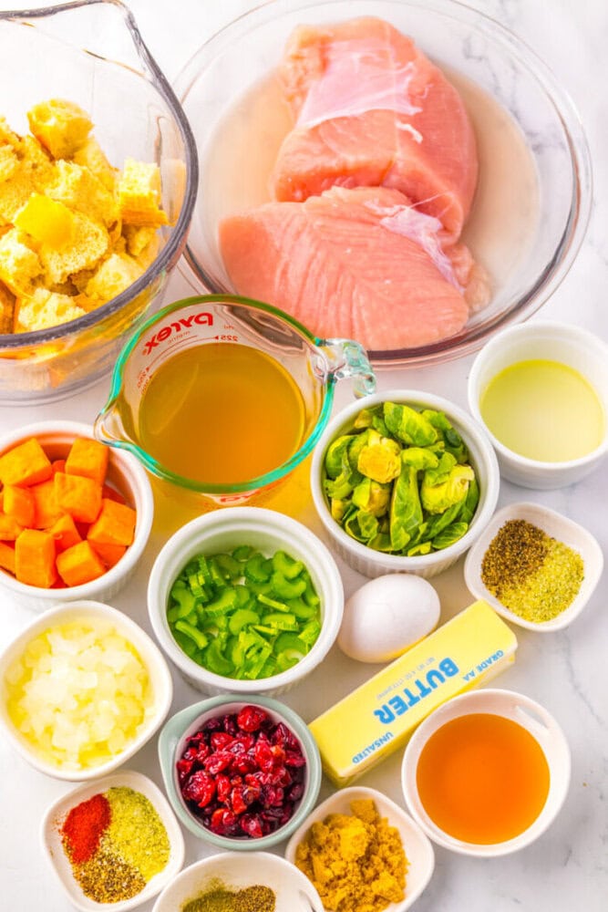 A collection of prepared ingredients for stuffing, including raw turkey breast, bread cubes, broth, vegetables, an egg, butter, cranberries, oil, and various seasonings on a white surface.