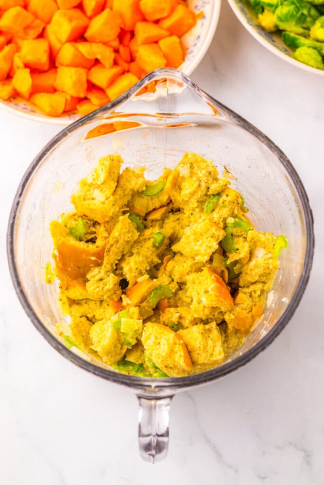A glass mixing bowl with bread stuffing mixture containing bread chunks and celery, with bowls of chopped carrots and broccoli in the background.