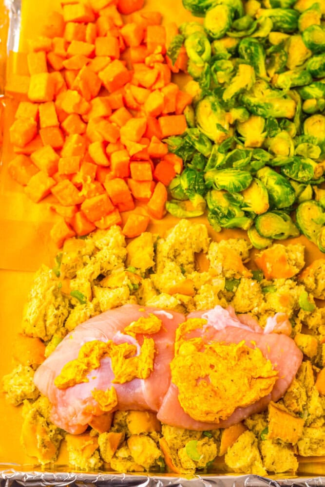Raw chicken breasts on top of seasoned stuffing cubes, with diced sweet potatoes and halved Brussels sprouts arranged on a foil-lined baking sheet.
