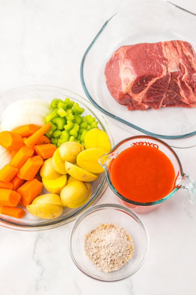 A glass dish with raw beef, a plate of sliced carrots, onions, celery, and potatoes, a measuring cup of tomato sauce, and a bowl of flour on a white surface.