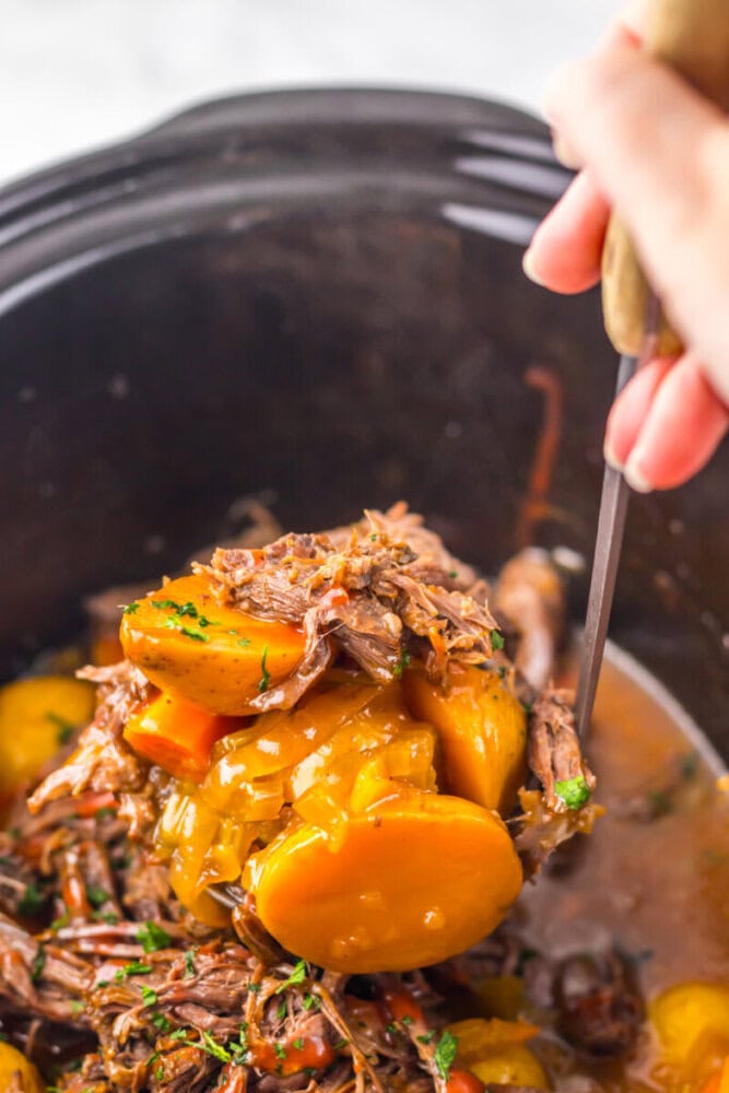 A hand lifts a serving of pot roast with potatoes, carrots, and gravy from a slow cooker using a large spoon.