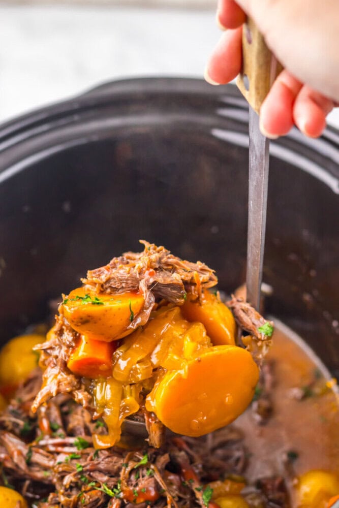 A hand holds a ladle filled with shredded beef, sliced carrots, potatoes, and gravy over a slow cooker.