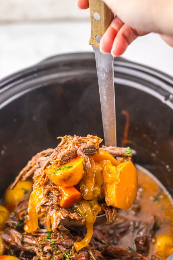 A hand uses a ladle to serve beef pot roast with carrots and potatoes from a slow cooker.