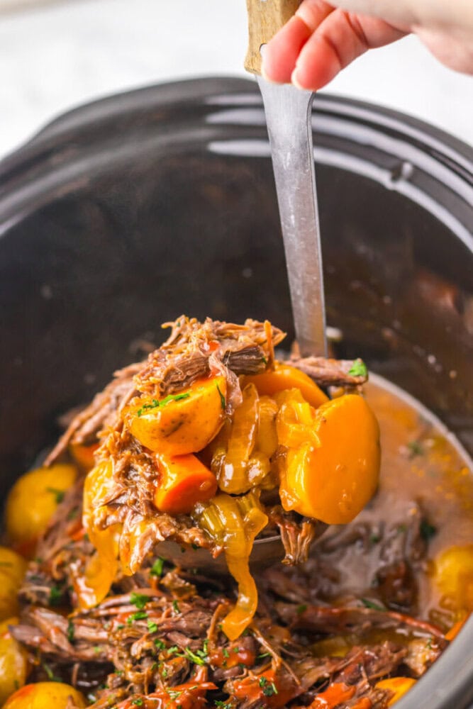 A ladle lifts pot roast with potatoes, carrots, and onions in a rich sauce from a slow cooker.