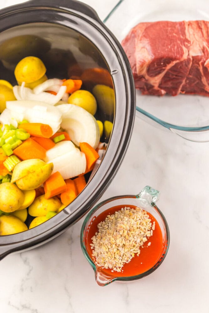 A slow cooker with chopped potatoes, carrots, onions, and celery, a glass dish with raw beef, and a measuring cup of tomato sauce topped with dried onions on a marble surface.
