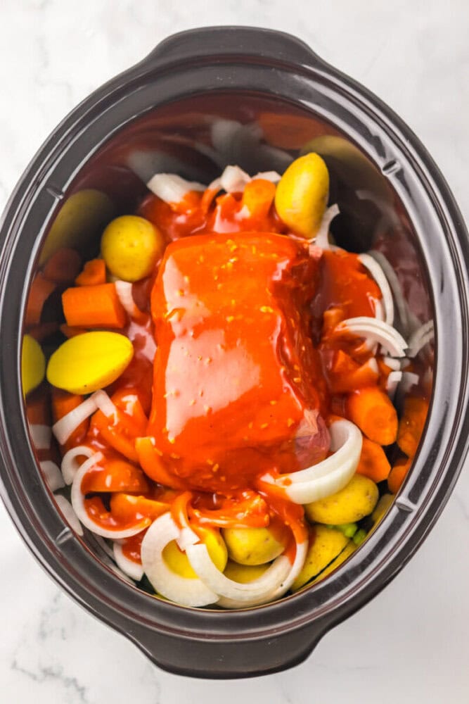 A slow cooker filled with potatoes, carrots, onions, and a piece of meat covered in red sauce, ready to be cooked.