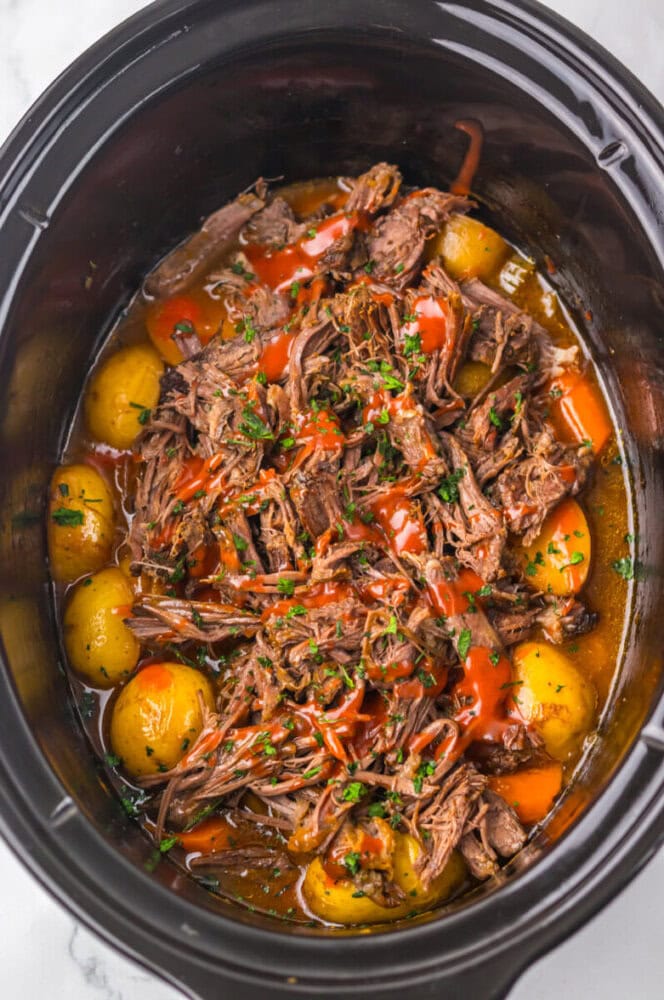 Shredded pot roast with baby potatoes in a slow cooker, topped with chopped herbs and a drizzle of sauce.