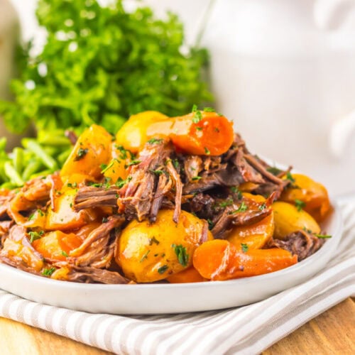 A plate of pot roast with shredded beef, potatoes, carrots, and green beans, garnished with parsley, sits on a striped napkin with fresh herbs in the background.