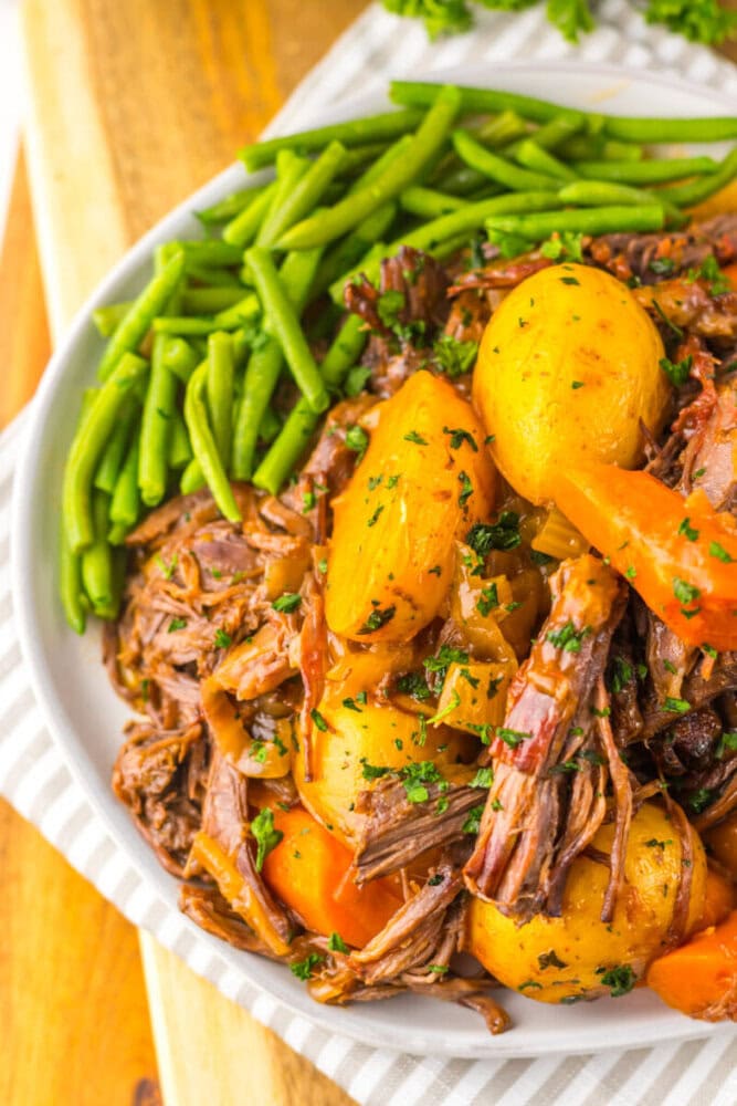 A plate of pot roast with shredded beef, potatoes, carrots, and green beans, garnished with chopped parsley.