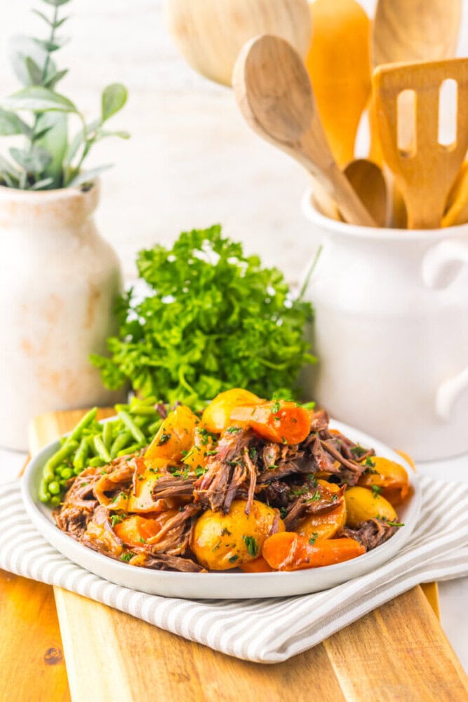 A plate of pot roast with shredded beef, potatoes, carrots, and green beans, garnished with parsley, sits on a striped cloth with kitchen utensils and a plant in the background.