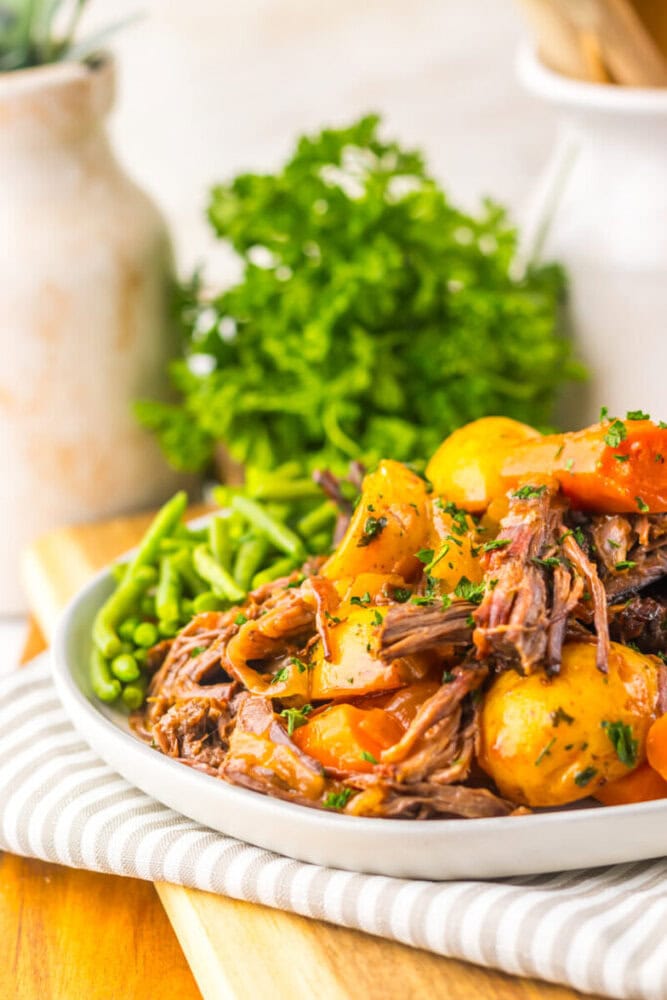 A plate of pot roast with shredded beef, potatoes, carrots, and green beans, garnished with herbs, sits on a striped napkin with fresh parsley in the background.
