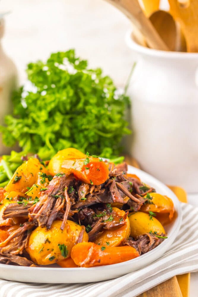 A plate of pot roast with shredded beef, baby potatoes, and carrots, garnished with chopped parsley, sits on a striped napkin with fresh herbs in the background.