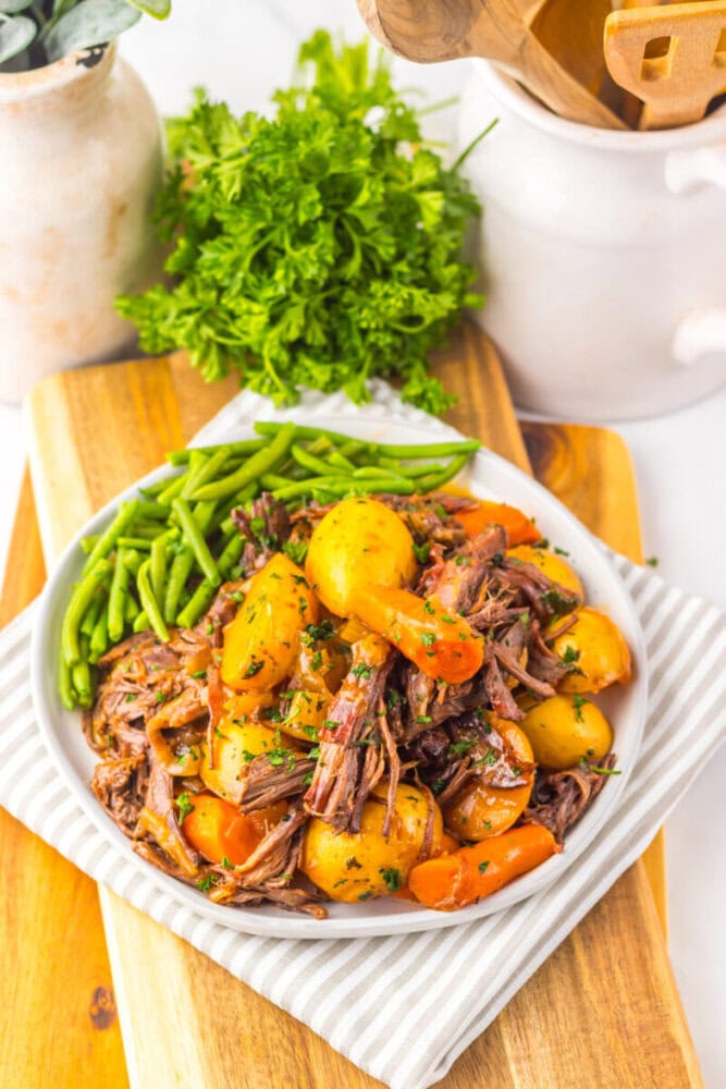 A plate of pot roast with shredded beef, baby potatoes, carrots, and green beans, garnished with parsley, sits on a wooden board next to a vase of fresh herbs.