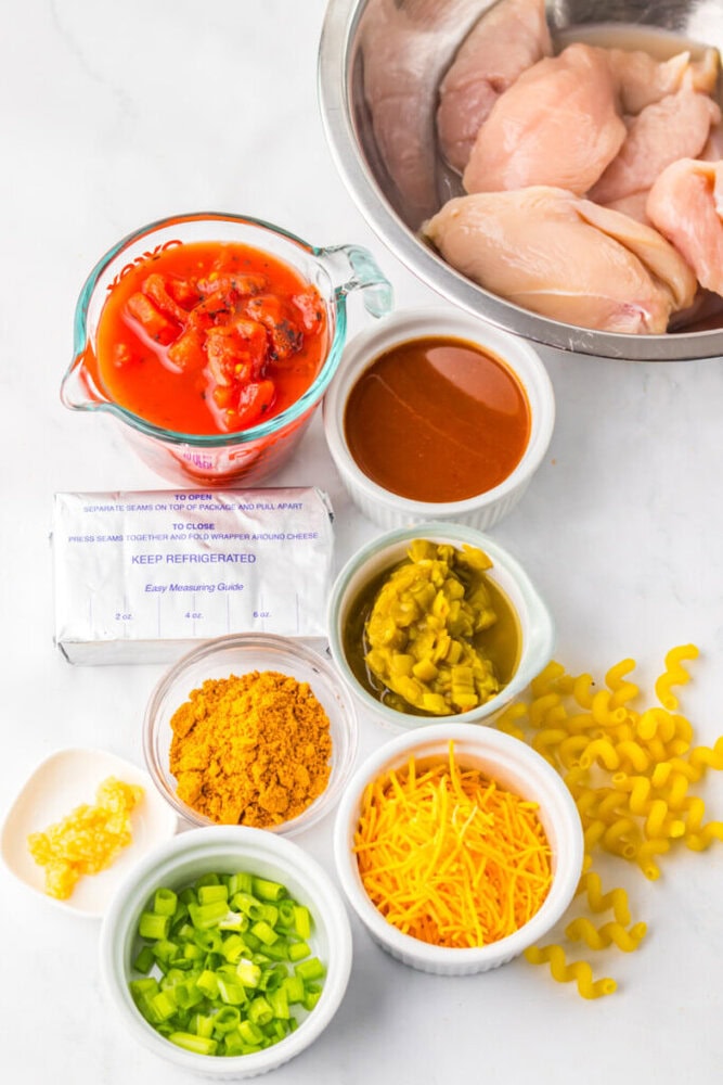 Ingredients for a recipe arranged on a countertop, including raw chicken breasts, diced tomatoes, sauce, cream cheese, green chiles, shredded cheese, curry powder, garlic, green onions, and dry pasta.