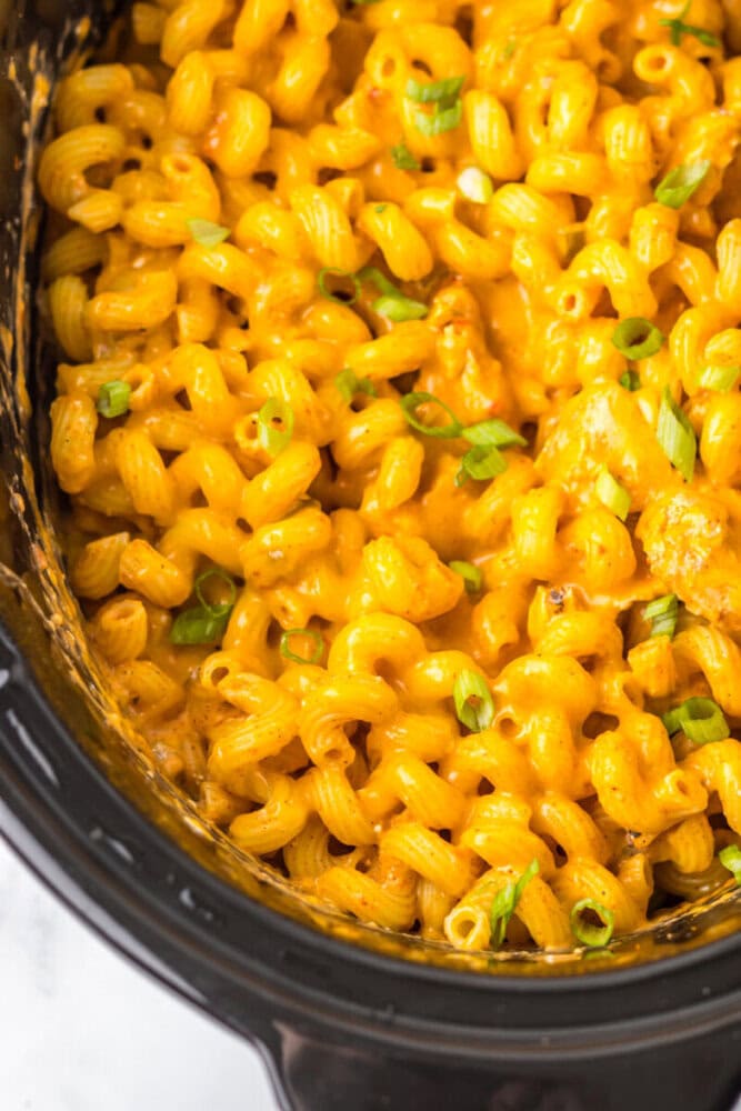 Cooked macaroni coated in creamy orange cheese sauce, topped with chopped green onions, inside a black slow cooker.