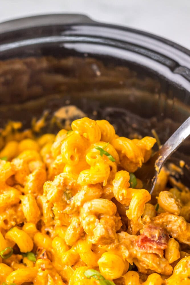 A close-up of cheesy macaroni and ground beef mixture being served from a black slow cooker with a metal spoon.