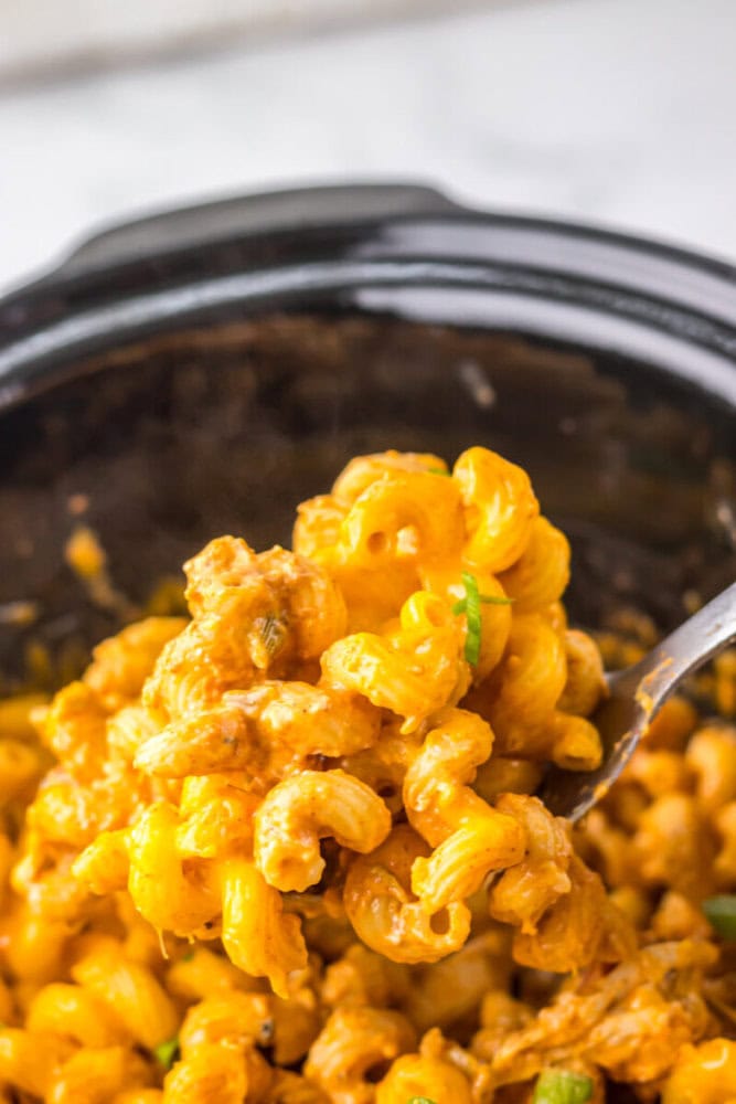 A close-up of a spoonful of cheesy pasta with ground meat and green peppers being lifted from a slow cooker.