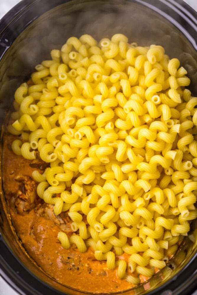 Uncooked cavatappi pasta sitting on top of a creamy tomato sauce with chicken in a slow cooker.
