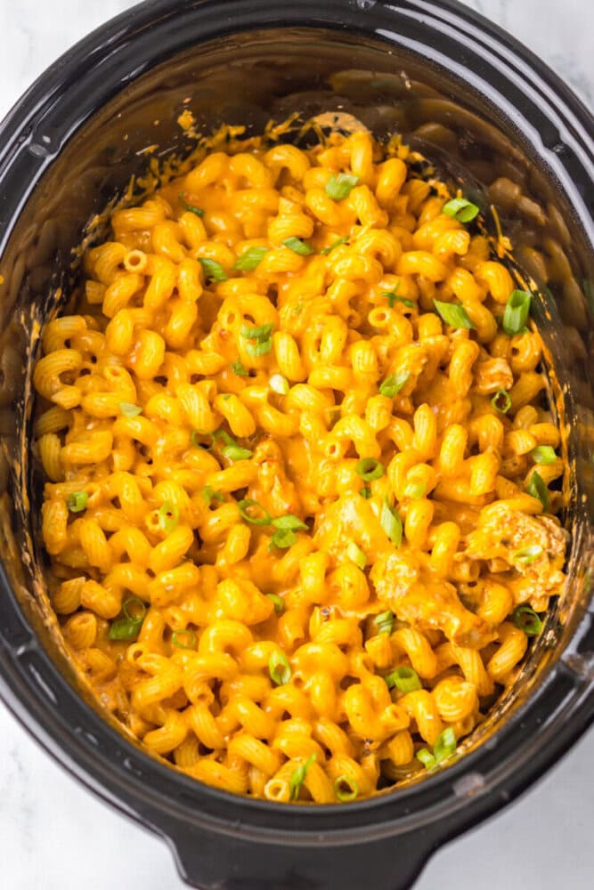 Cooked cavatappi pasta mixed with melted cheese and topped with chopped green onions in a black slow cooker.