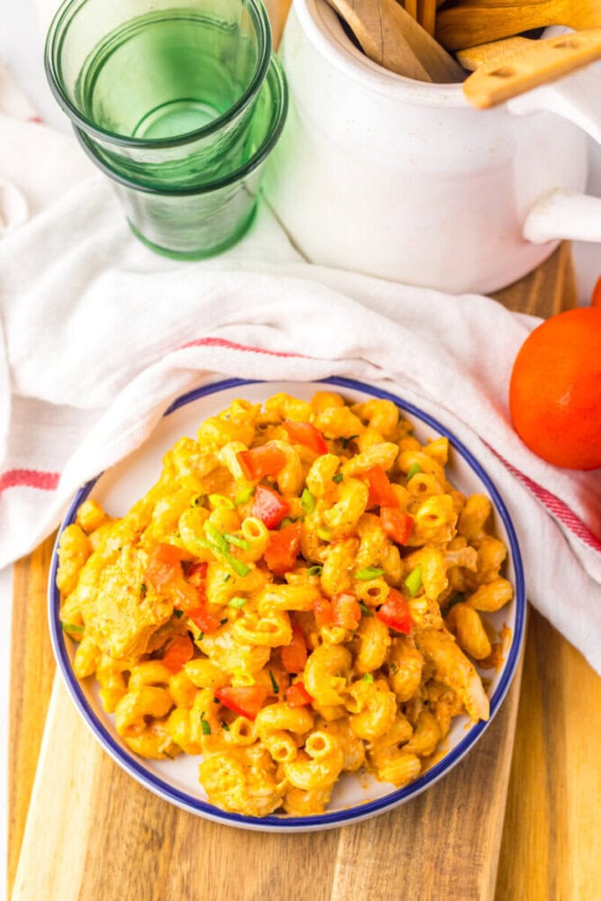 A plate of macaroni pasta mixed with diced red bell peppers and a creamy orange sauce, placed on a wooden surface next to a tomato, glass cups, and kitchen utensils.