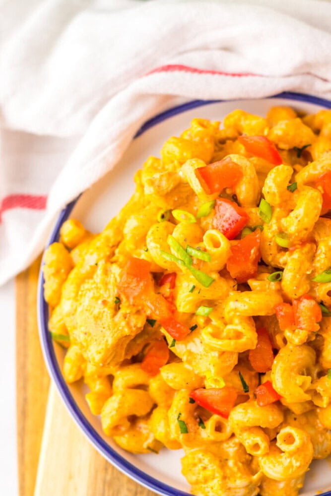 A plate of macaroni pasta mixed with a creamy orange sauce, diced red bell peppers, and chopped green herbs, placed next to a white cloth napkin.