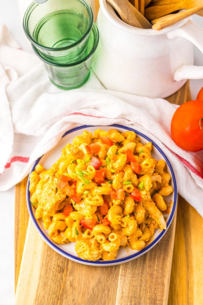 A plate of macaroni pasta mixed with diced red bell peppers and sauce sits on a wooden board next to green glasses, a white teapot, and fresh tomatoes.