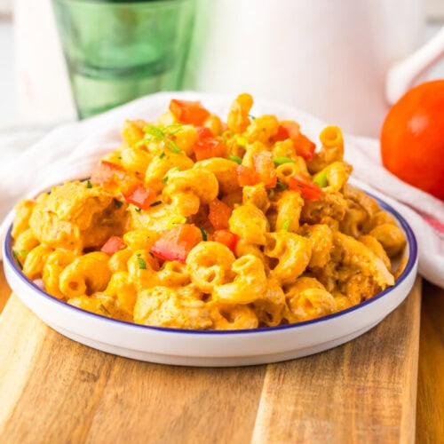 A plate of macaroni pasta with creamy orange sauce, diced tomatoes, and herbs, placed on a wooden board with whole tomatoes and a green glass in the background.
