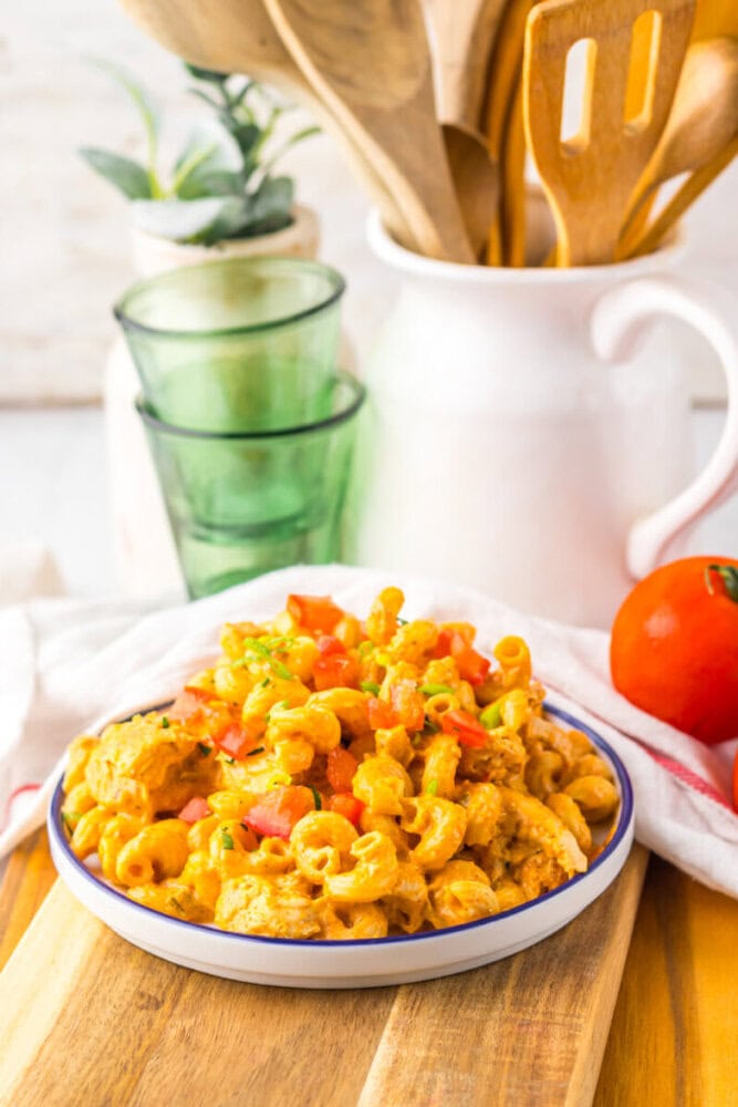 A plate of macaroni pasta in a creamy orange sauce, topped with diced tomatoes and herbs, sits on a wooden board with kitchen utensils, glasses, and a tomato in the background.