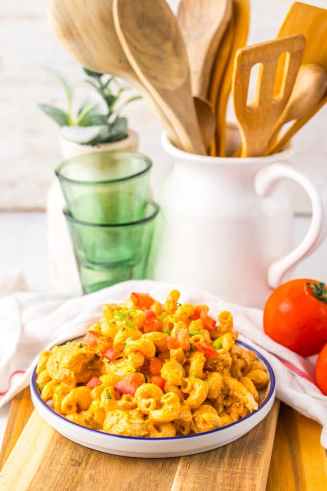A plate of macaroni and cheese topped with diced tomatoes and green onions sits on a wooden board; kitchen utensils and tomatoes are in the background.