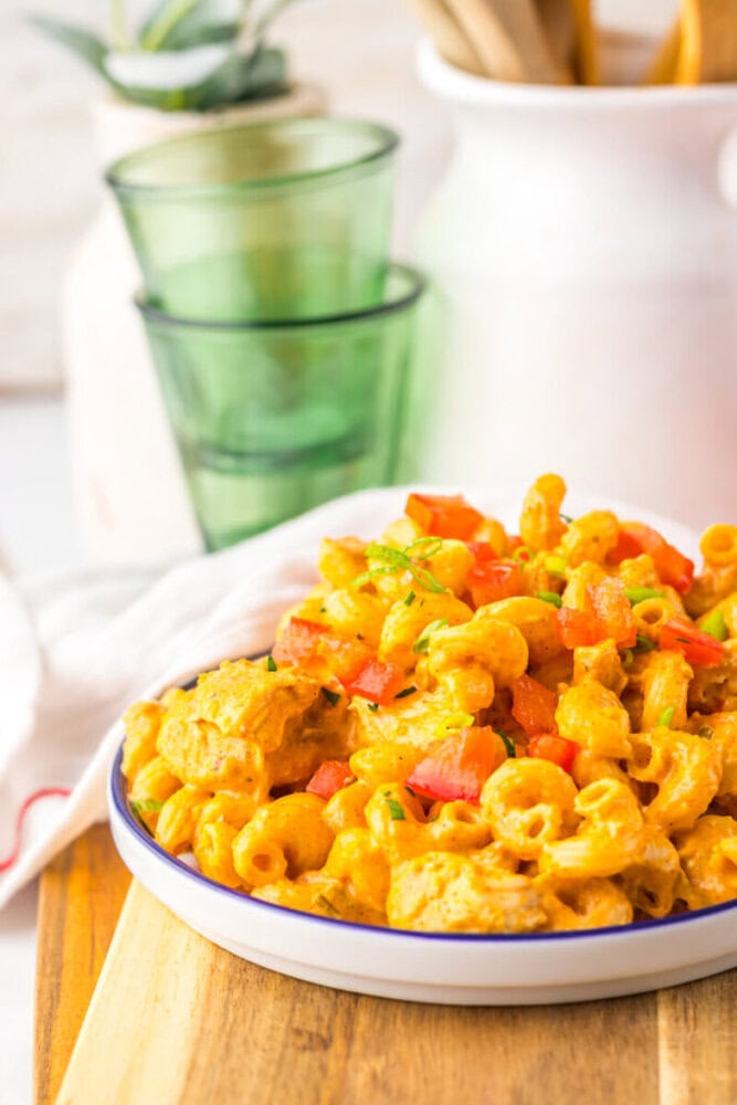 A plate of macaroni pasta mixed with diced tomatoes and herbs sits on a wooden board, with green glasses and kitchen items in the background.