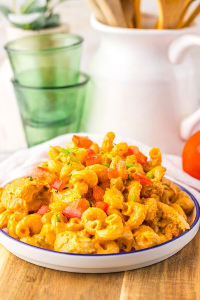 A plate of creamy macaroni pasta mixed with diced tomatoes and green herbs, served on a wooden table with glasses and kitchen utensils in the background.
