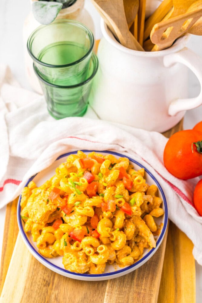 A plate of pasta with a creamy orange sauce and diced vegetables sits on a wooden board next to green glasses, a white utensil holder, and two orange fruits.