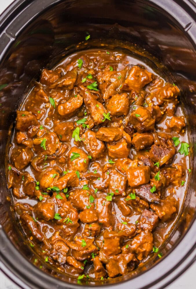 Chunks of beef stew in a rich brown sauce, garnished with chopped parsley, inside a black slow cooker.