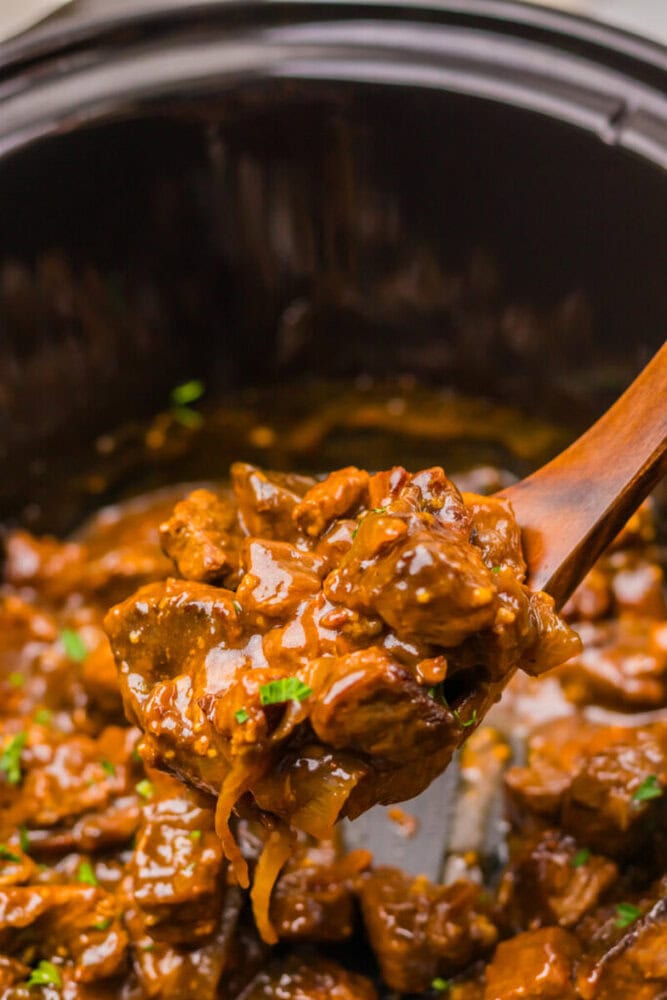 A wooden spoon lifts saucy beef stew with onions and herbs from a slow cooker.
