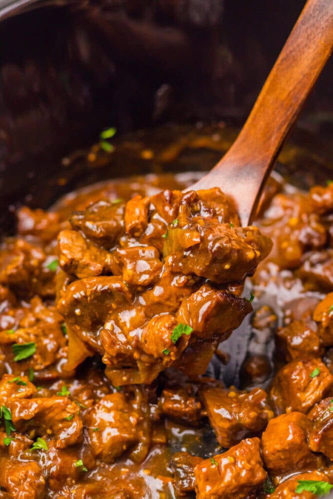 Close-up of a wooden spoon scooping saucy, cooked beef stew with small pieces of meat and herbs in a dark pot.