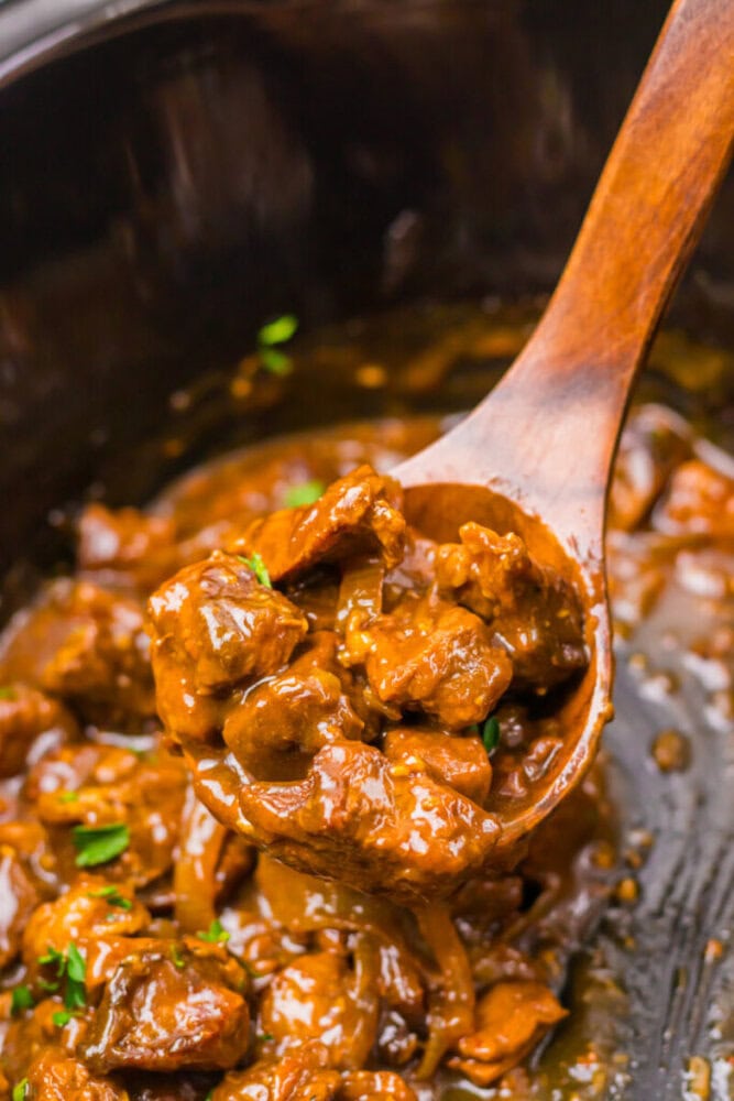 A wooden spoon lifts a serving of beef stew with gravy and herbs from a slow cooker.