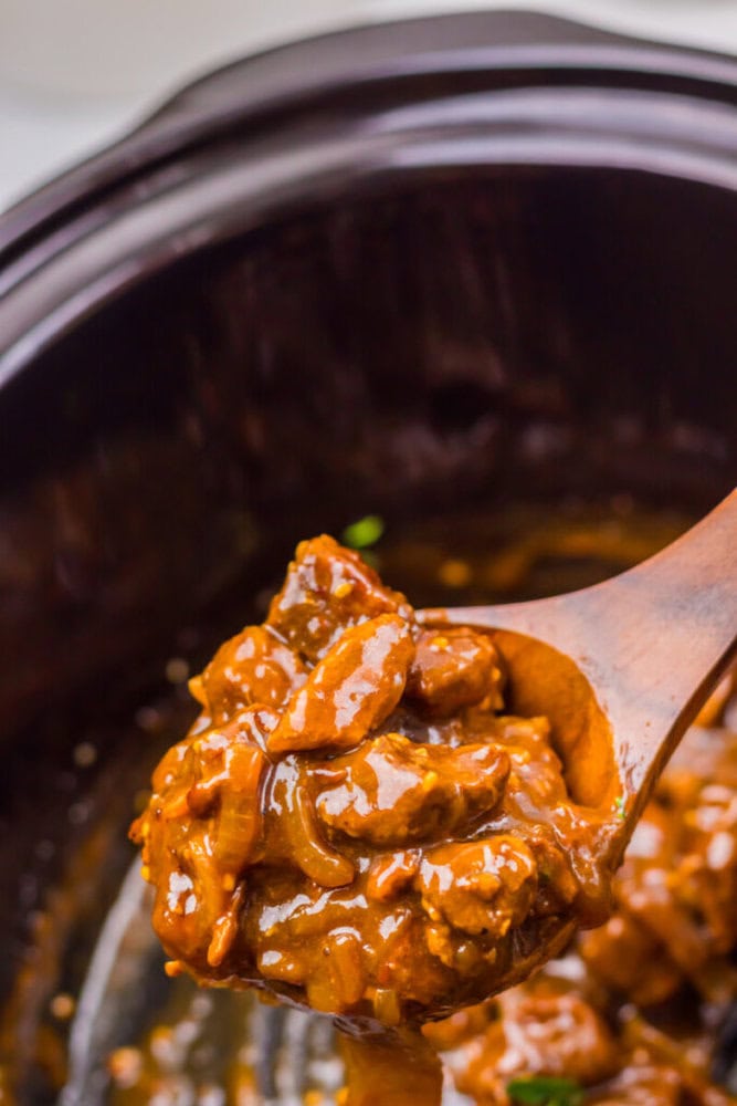 A wooden spoon holds a serving of thick, saucy beef stew with visible chunks of meat and beans inside a slow cooker.