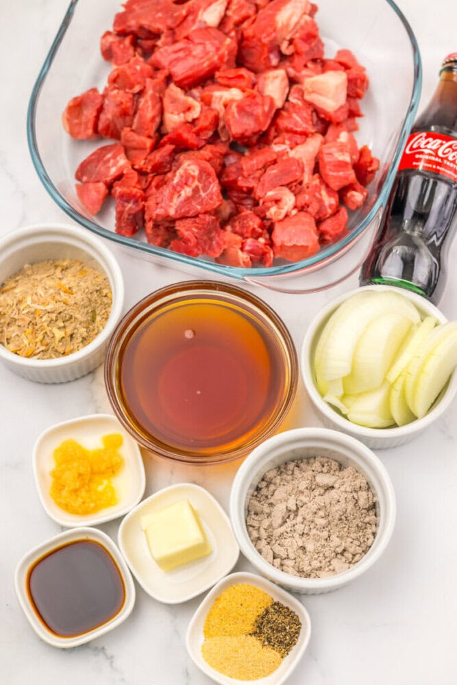 A glass bowl of cubed raw beef, a bottle of Coca-Cola, sliced onions, broth, butter, dry soup mix, garlic, soy sauce, seasoning mix, and spices on a white surface.