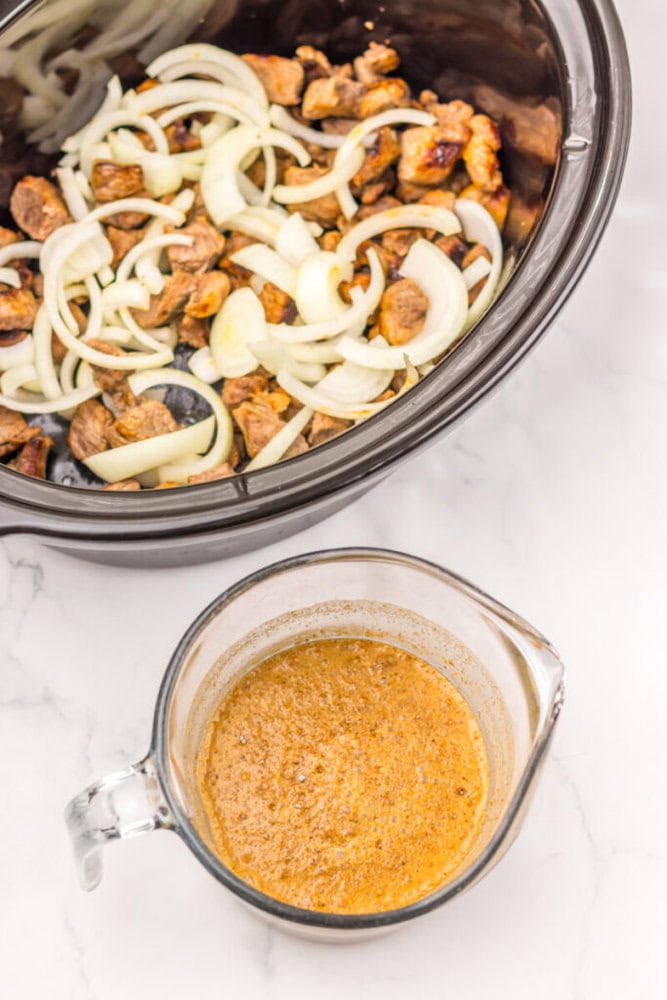 Sliced onions and pieces of meat in a slow cooker, with a measuring cup of seasoned liquid mixture on a marble countertop nearby.