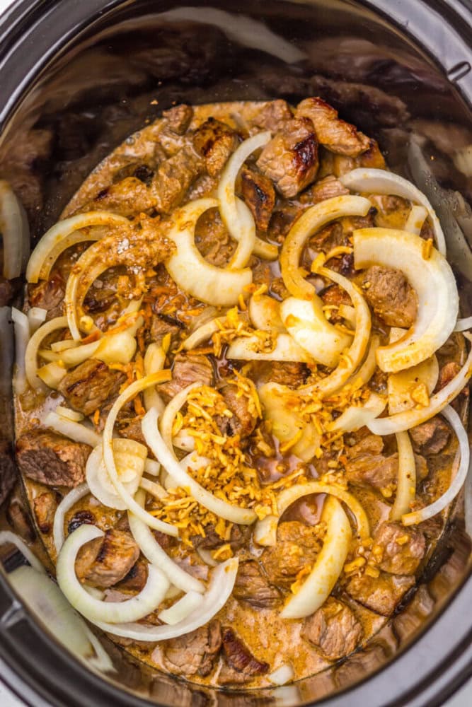 Sliced onions and browned beef pieces with seasonings in a slow cooker, ready to be cooked.
