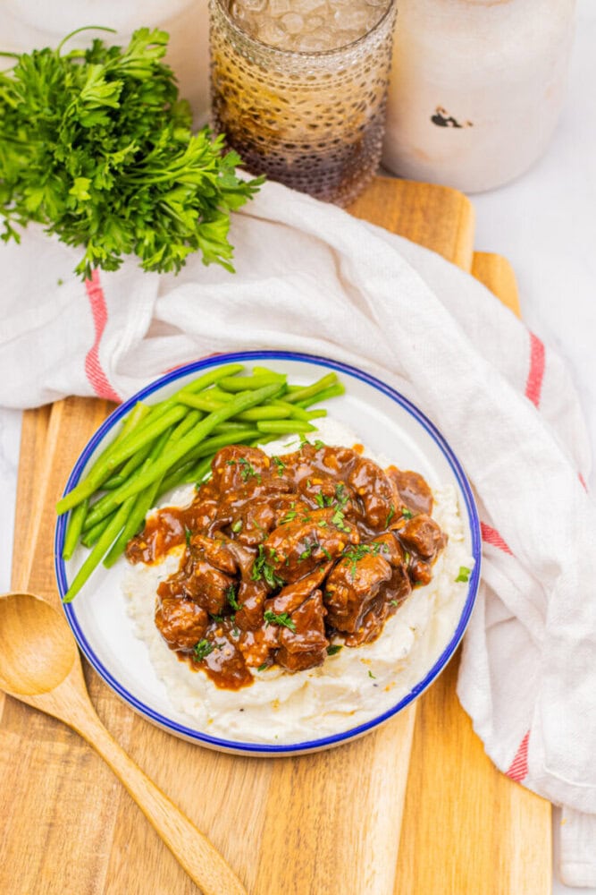 A plate of beef stew served over mashed potatoes with green beans on the side, placed on a wooden board with a wooden spoon, parsley, and a drink nearby.